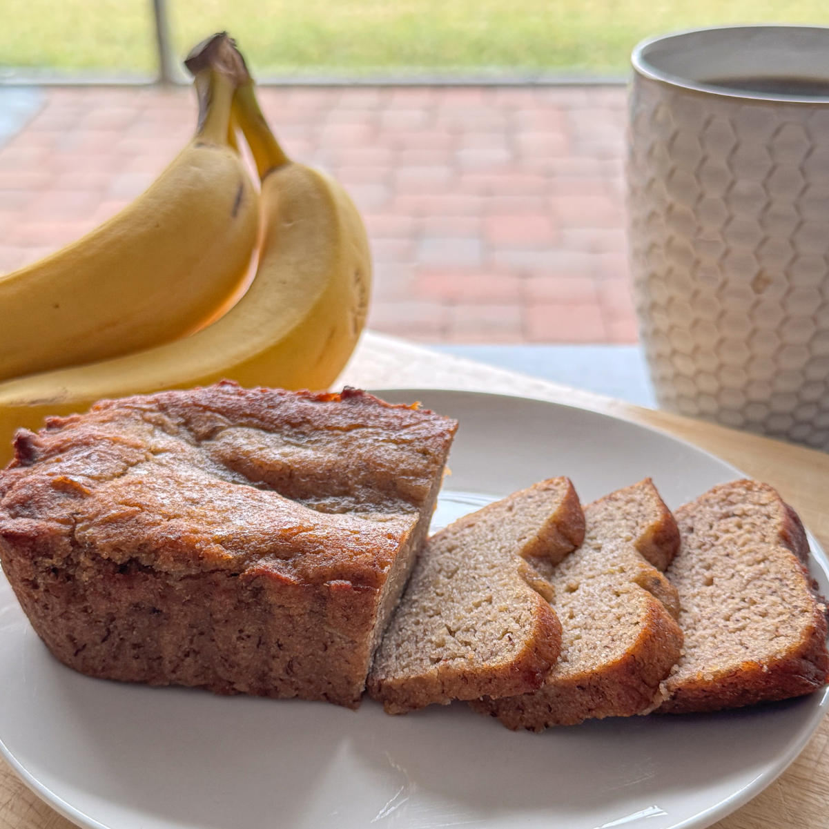 Cozy lifestyle shot of freshly sliced banana bread on a white plate, accompanied by ripe bananas and a warm cup of coffee. The outdoor patio setting adds a relaxed, inviting vibe, perfect for showcasing a comforting snack or breakfast moment