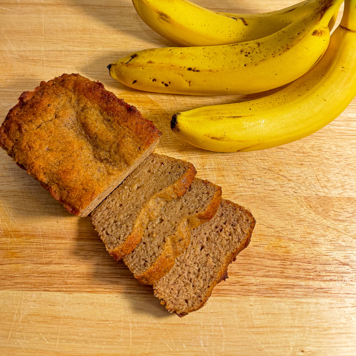 Freshly baked banana bread loaf, sliced to reveal its moist and fluffy texture, paired with ripe bananas on a wooden cutting board. Perfect for showcasing natural ingredients and homemade quality